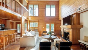 A spacious living area with a high ceiling, wooden walls, a fireplace, and a kitchen area with a bar. - Raven by Avantstay Entertainers Dream w/ Pool, Hot Tub, Game Room! 9 Brs! (Sullivan's Island)