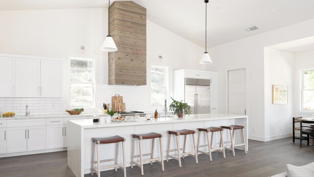 Une cuisine moderne avec des meubles blancs, un îlot central doté de tabourets, un éclairage suspendu et un mur d’accent en bois.