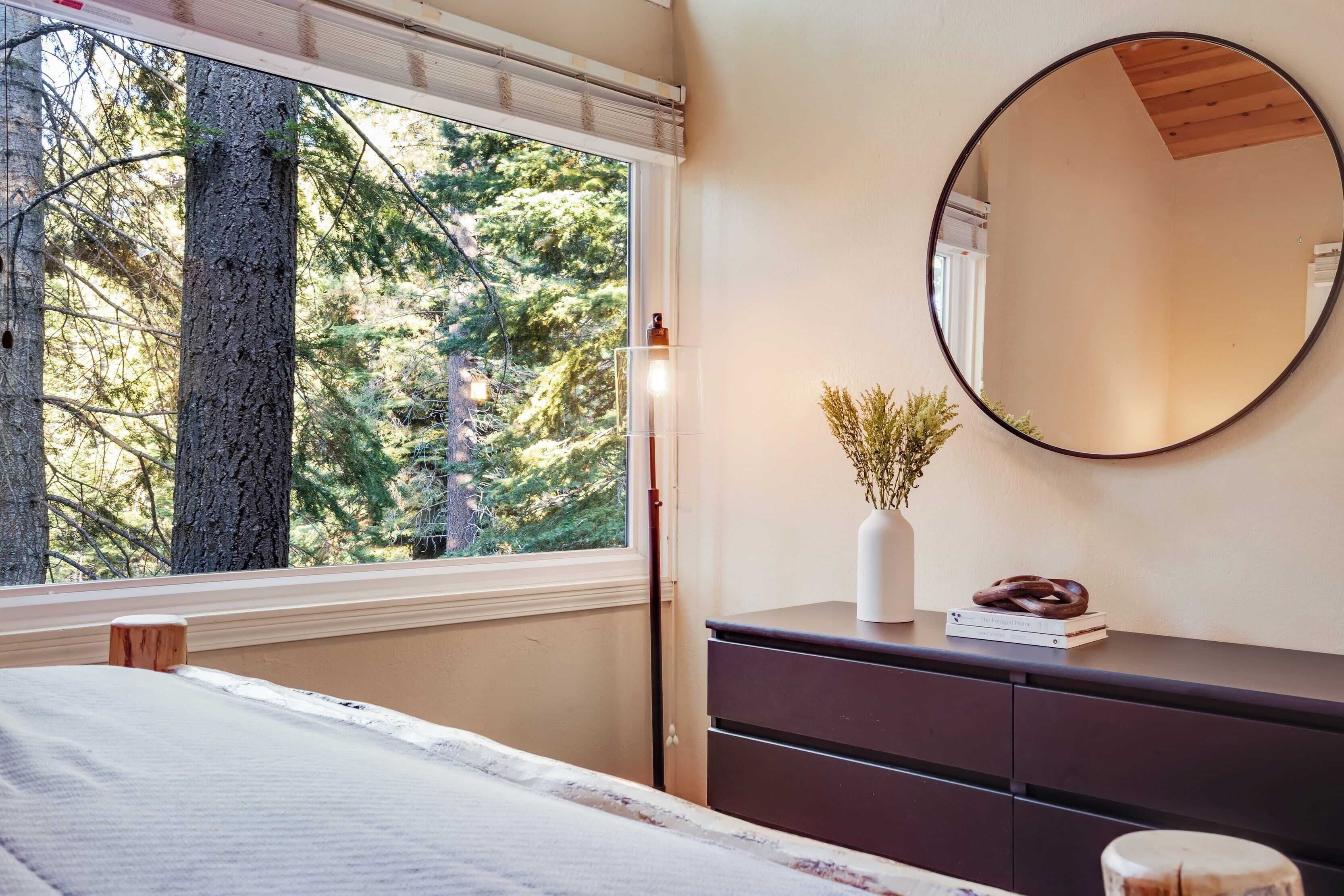 A bedroom with a large window, a bedside table with a vase and books, and a dresser.