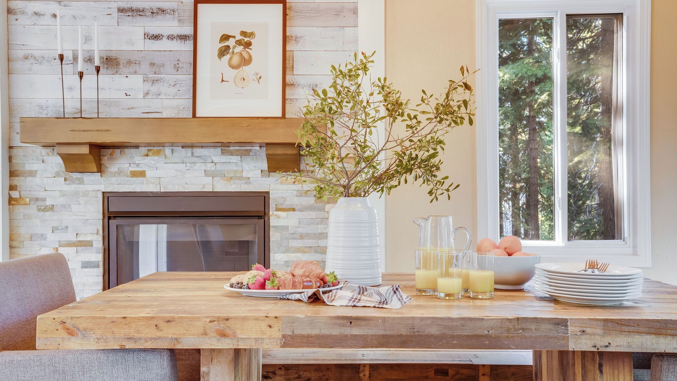 A dining area with a wooden table, a fireplace, a vase with branches, and a window with a view of trees.