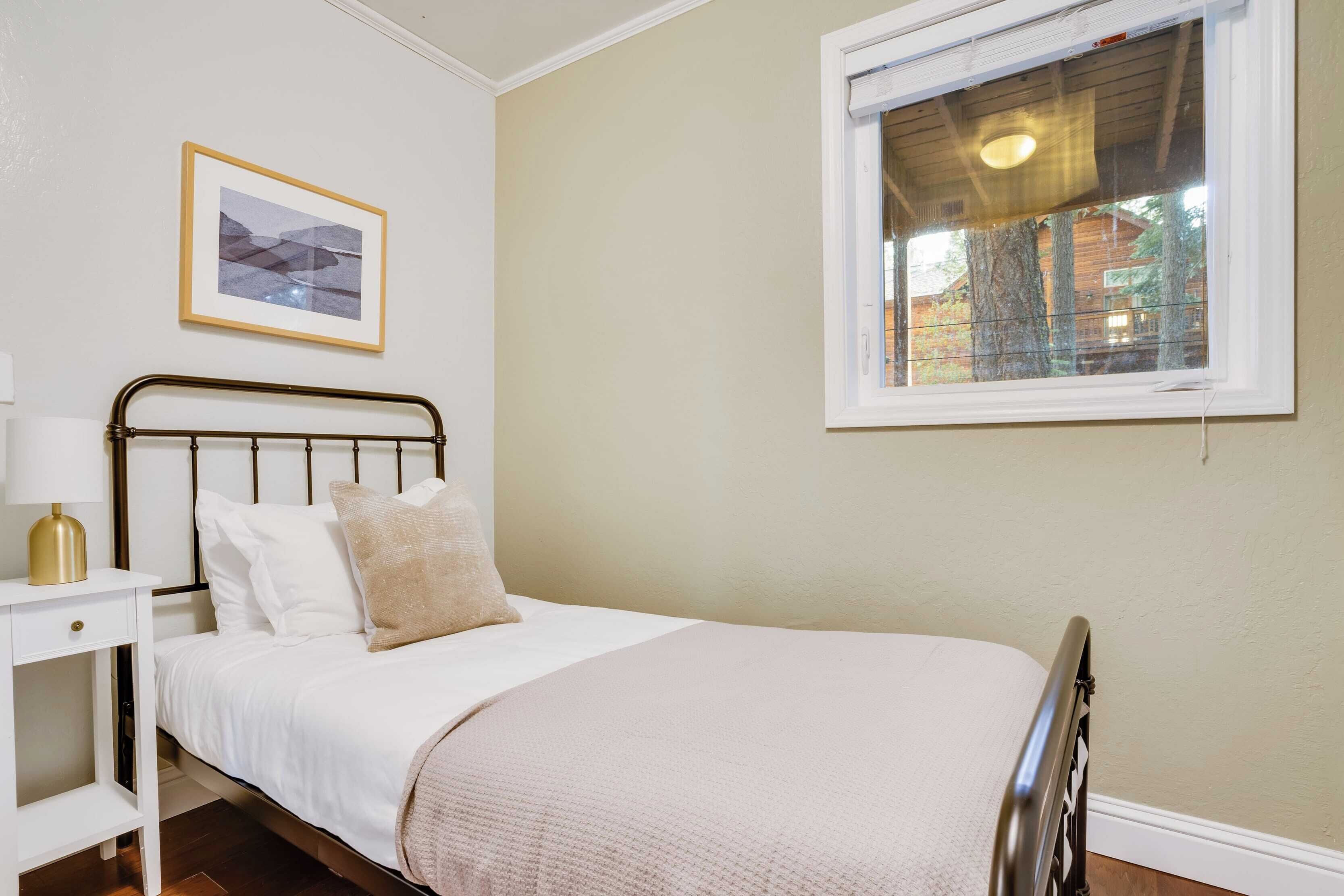 Un dormitorio ordenado con cama, mesita de noche, lámpara, cuadro enmarco y ventana con vistas a árboles y un edificio.