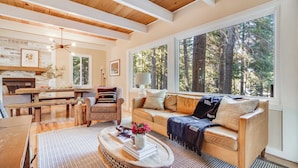 A cozy living room with a wooden ceiling, large windows offering a forest view, a sofa with cushions, a coffee table with books and a vase, and a fireplace. - Hot Tub, Sauna, Game Room Modern 5bdr Family Cabin Homewood by Avantstay (Homewood)