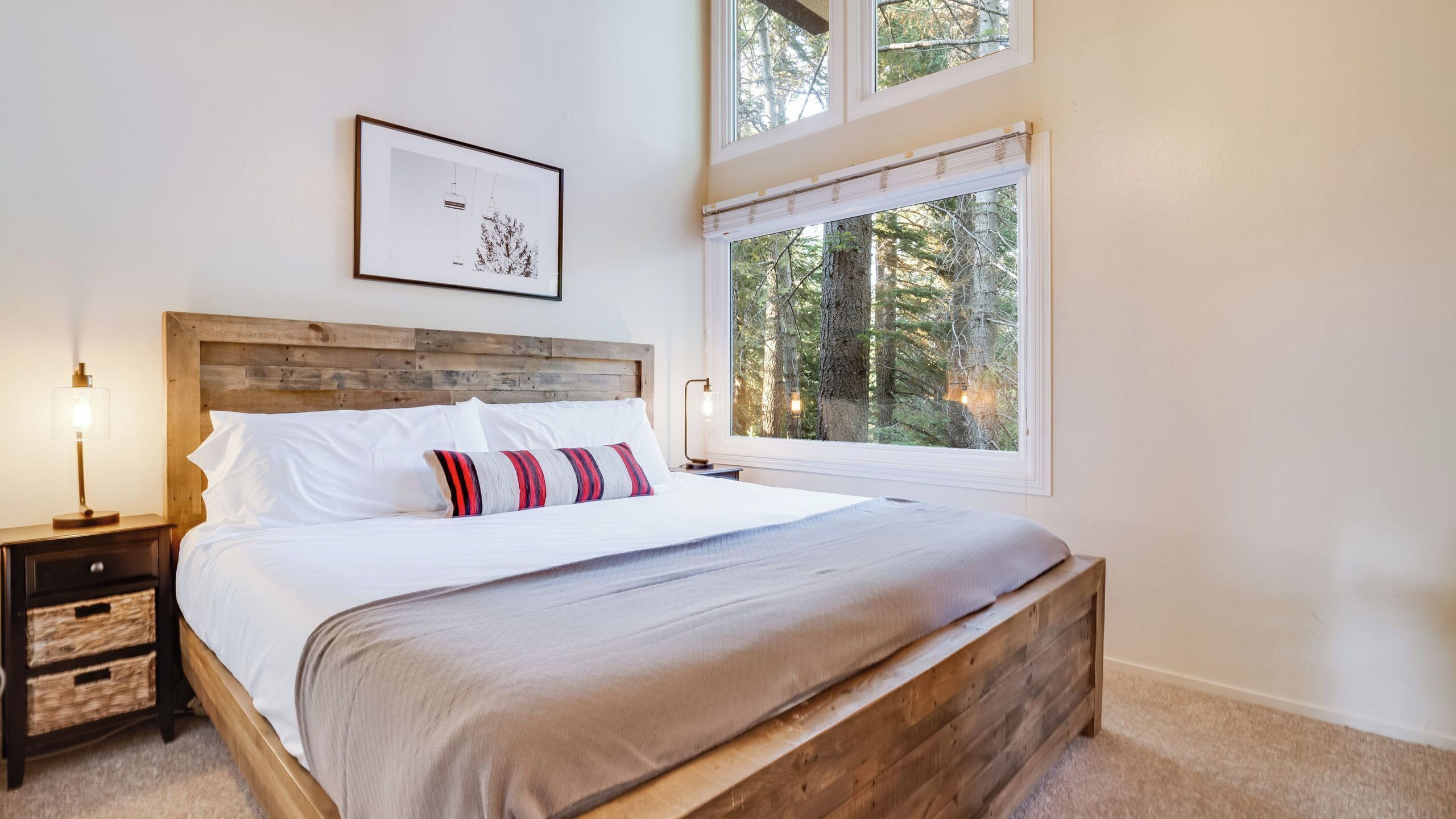 Een slaapkamer met een houten hoofdeinde, een bed met wit beddengoed en een gestreept kussen, een nachtkastje met een lamp en een raam met uitzicht op bomen.
