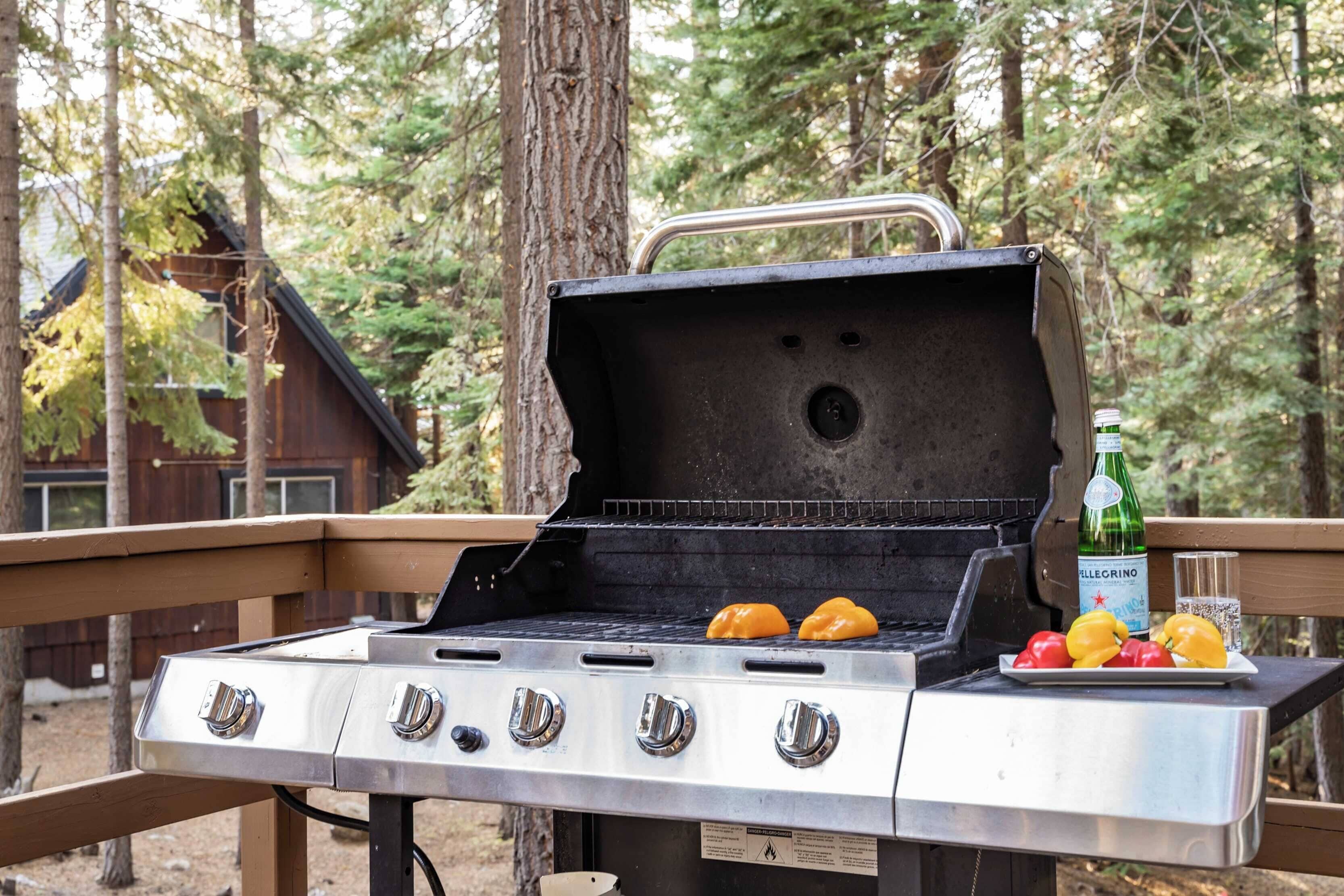 A portable grill with four burners and a metal handle, placed outdoors with a wooden deck and trees in the background.