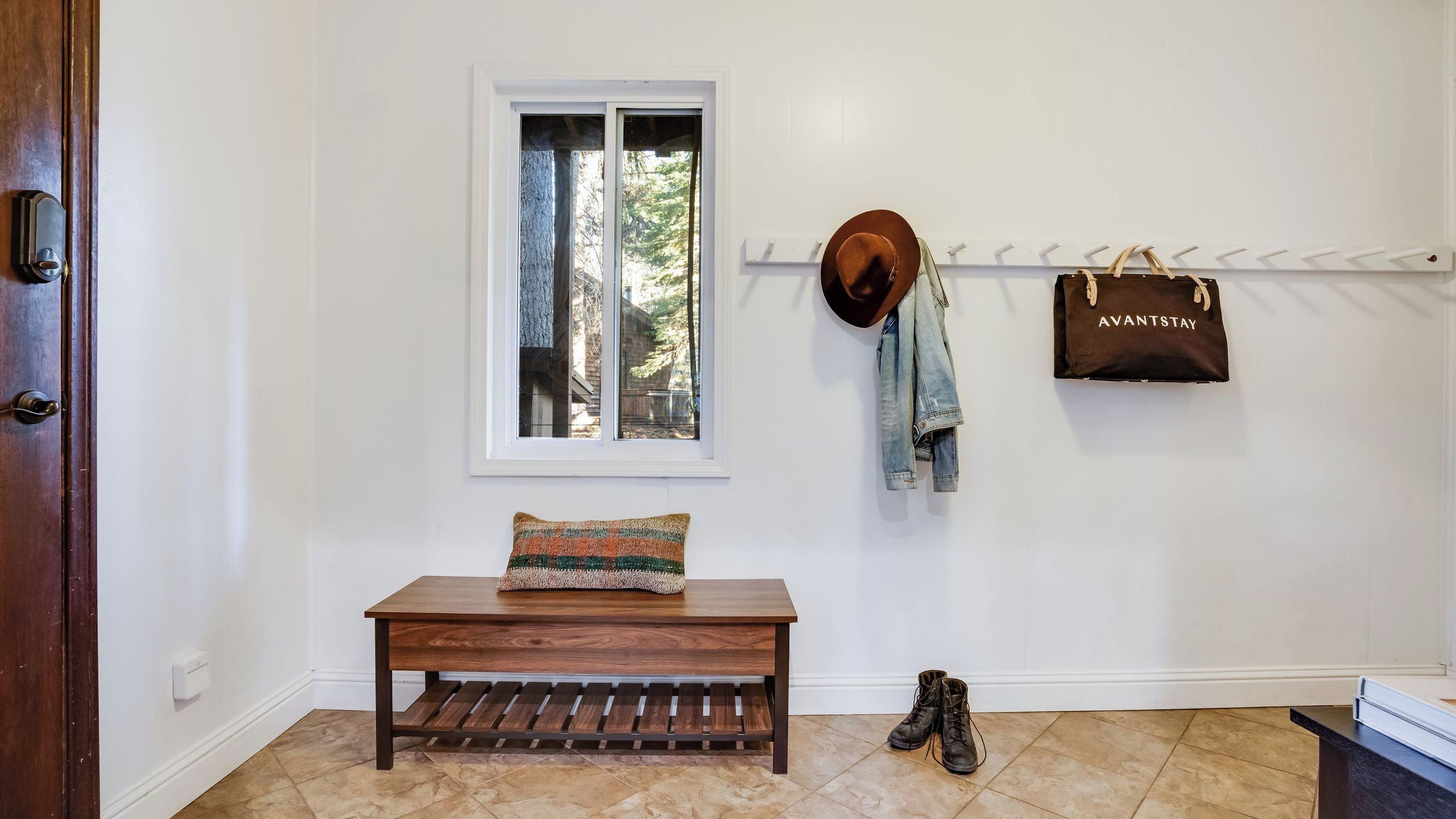 A wooden bench with a patterned cushion, a window with a view of trees, a hat, a denim jacket, and a brown bag hanging on a rack.