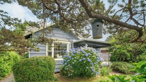 A house with a large window, a birdhouse, and a bush with blue flowers. - Beach Pines Bungalow by Avantstay Classic Cottage Steps to the Beach w/ Private Yard (Arch Cape)