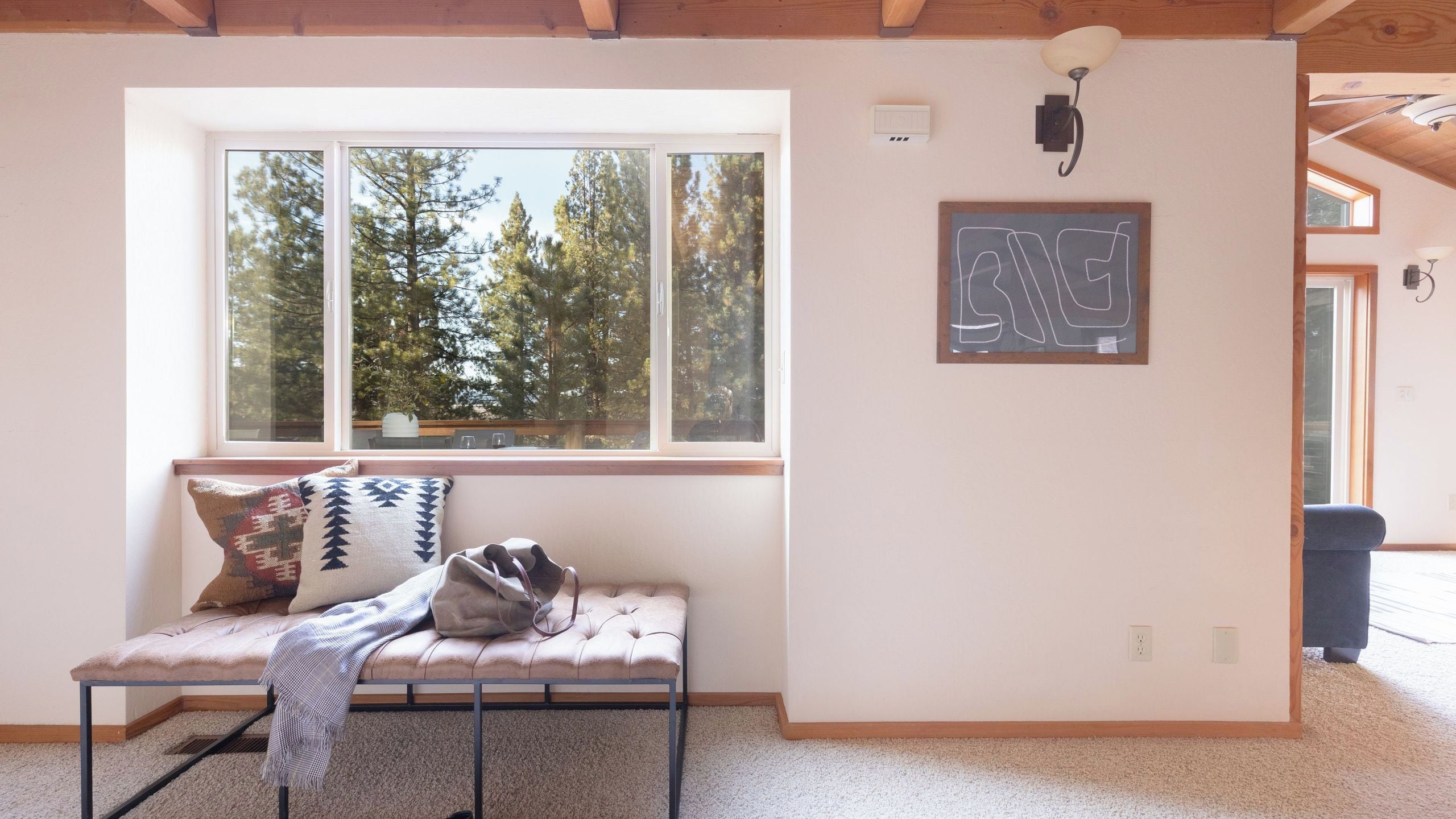 A room with a bench, a window with a view of trees, a wall-mounted light fixture, and a framed word on the wall.