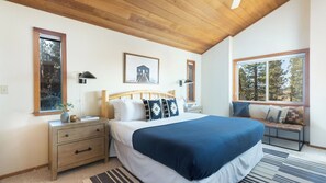 A bedroom with a wooden ceiling, a bed with a blue blanket, a nightstand, a bench by the window, and a framed picture on the wall.