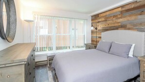 Een slaapkamer met een bed, een dressoir, een nachtkastje, een lamp en een raam met jaloezieën.