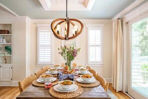 A dining room with a wooden table set for a meal, surrounded by chairs. A chandelier hangs from the ceiling, and there are windows with curtains on the walls.