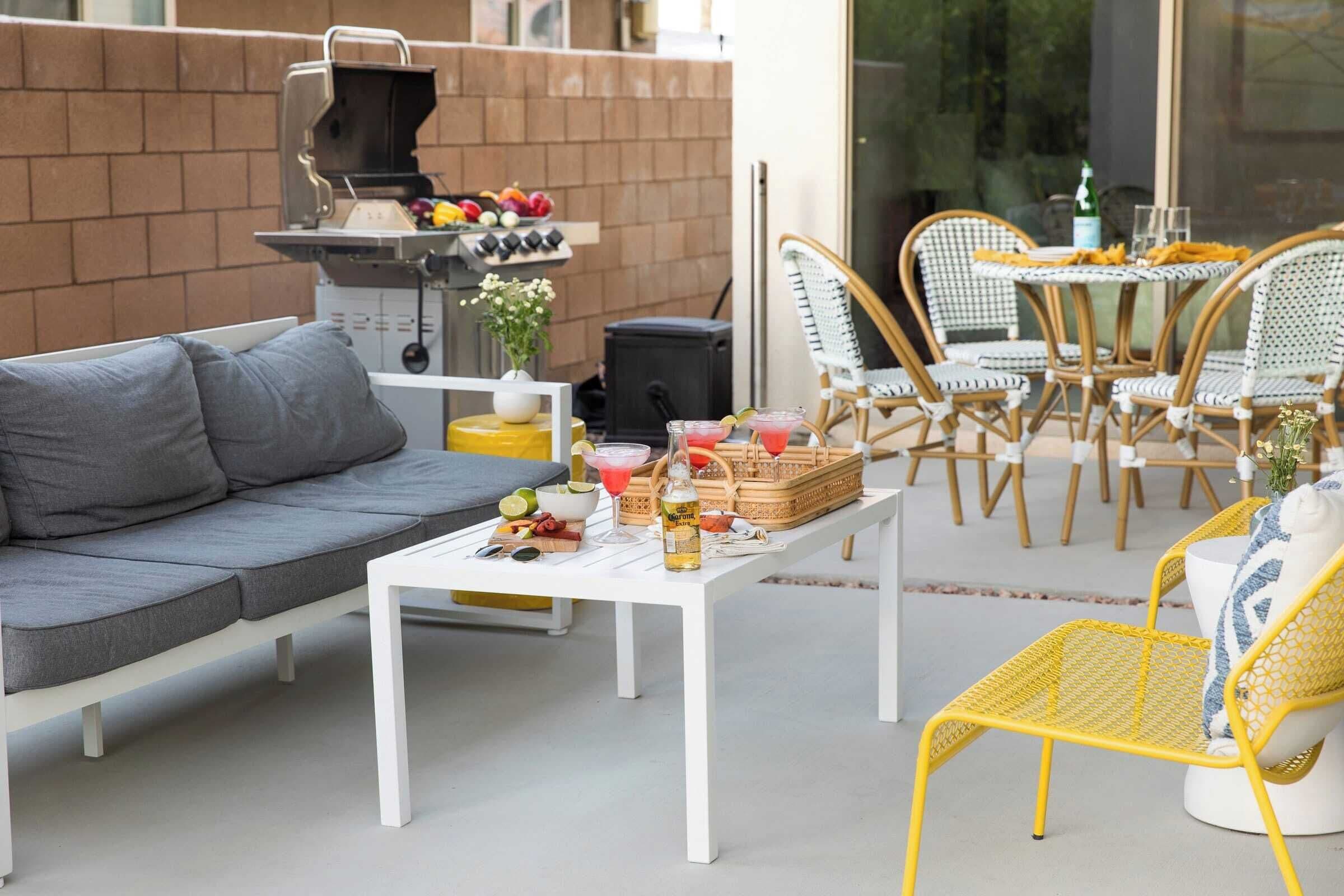 Un patio al aire libre con un sofá blanco, sillas amarillas, una mesa blanca con bebidas y aperitivos, una parrilla y sillas de mimbre.