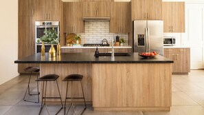 A modern kitchen with wooden cabinets, stainless steel appliances, a black granite countertop, and a central island with stools. - Marilyn by Avantstay Mins to Old Town La Quinta! (La Quinta)