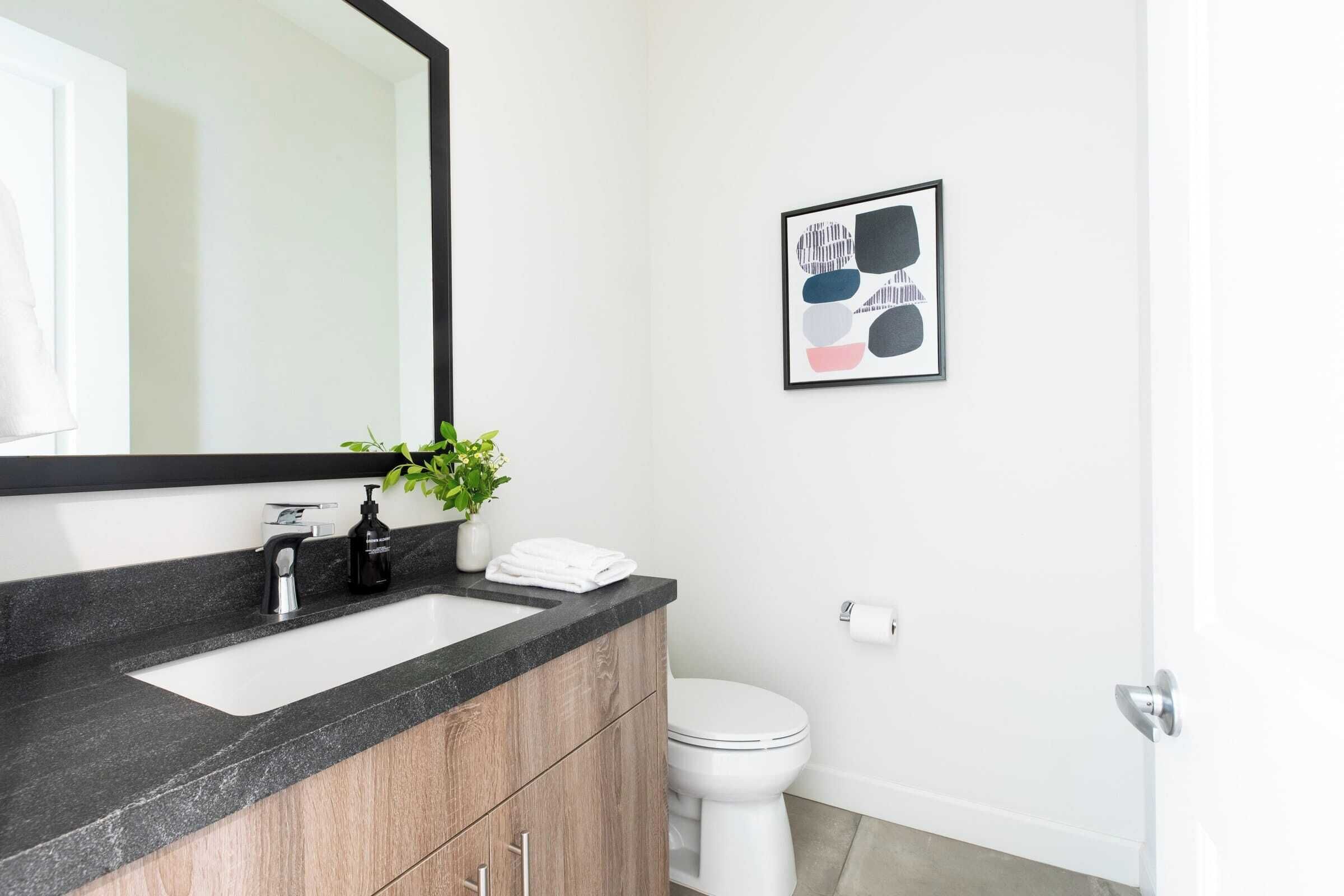 A modern bathroom with a black countertop sink, wooden cabinets, a white toilet, and a framed abstract art piece on the wall.