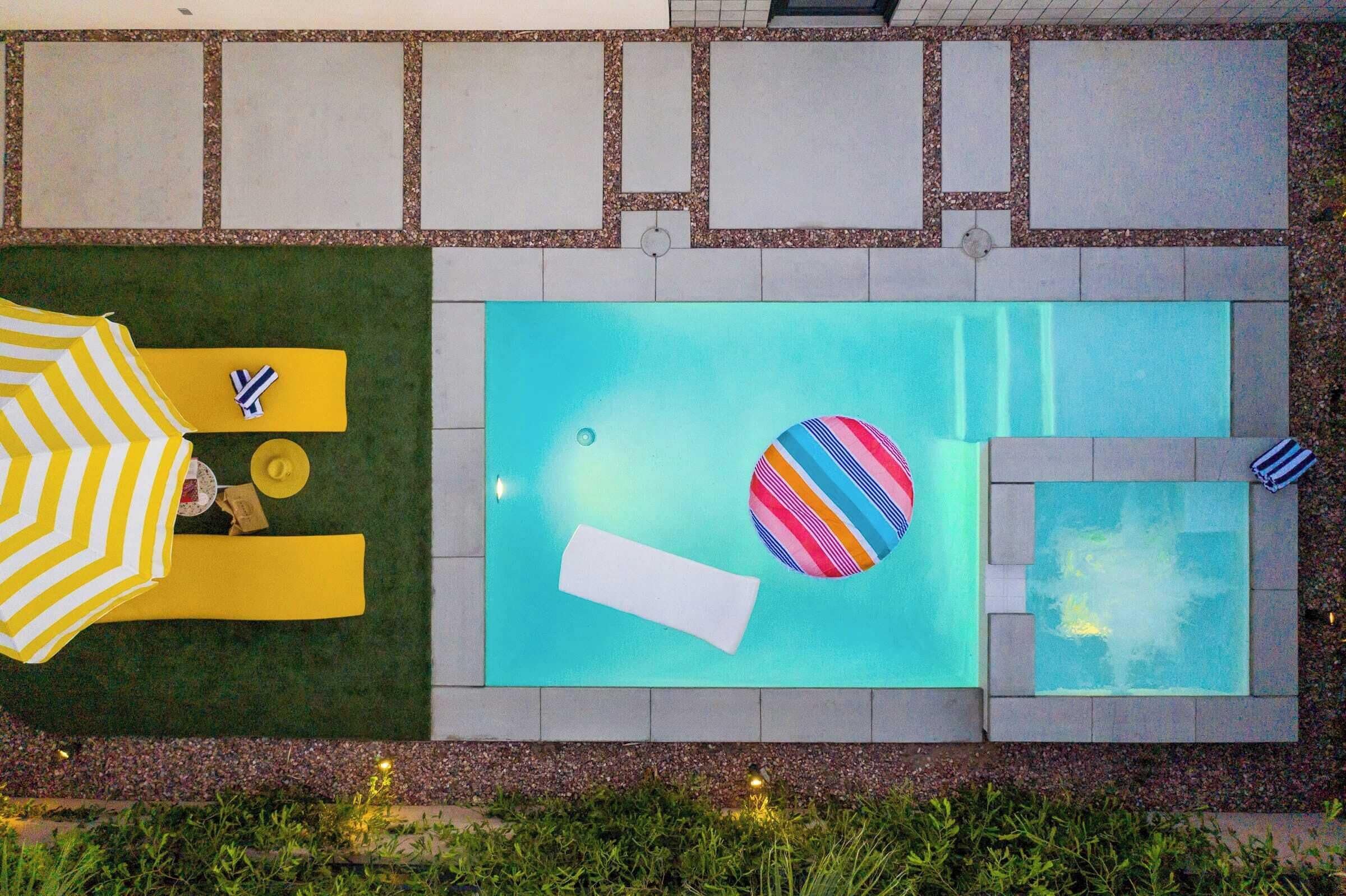 A rectangular swimming pool with a tiled deck, a yellow and white striped umbrella, and a colorful striped ball.