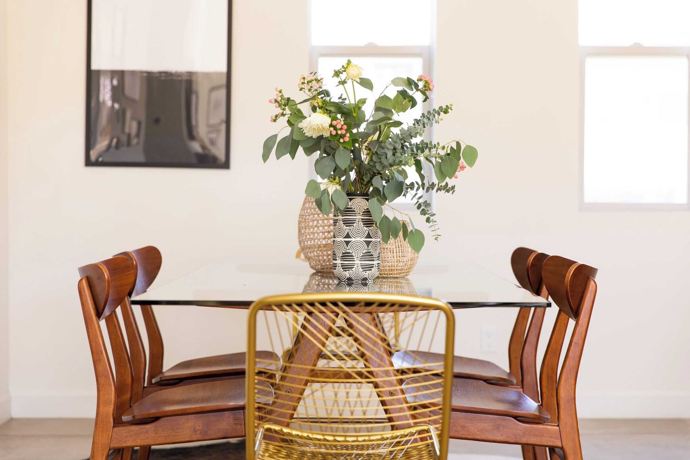 A dining table with a glass top and two wooden chairs with woven backs. A decorative vase with flowers is placed on the table.