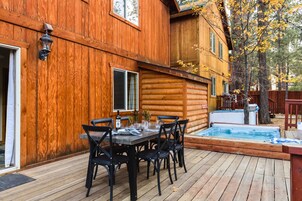 A wooden cabin with a deck, a hot tub, and a dining area with chairs and a table.