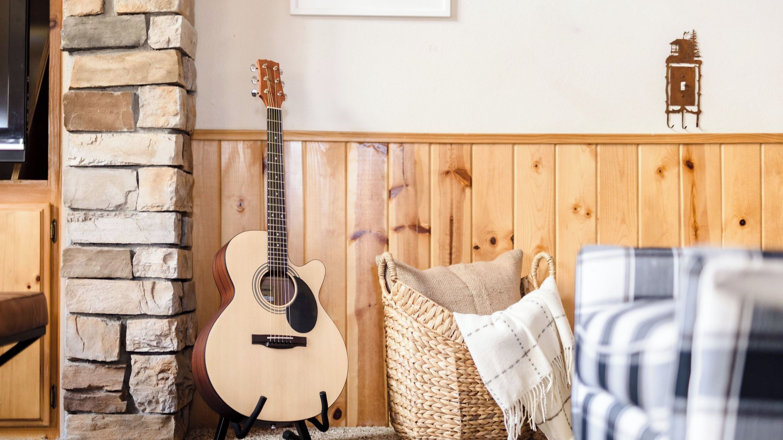 Une pièce avec un mur en pierre, des lambris en bois, une guitare, un panier tressé et une couverture à carreaux.
