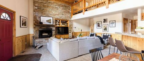 A cozy living room with a stone fireplace, a grey sofa, a wooden dining table, and a red door.