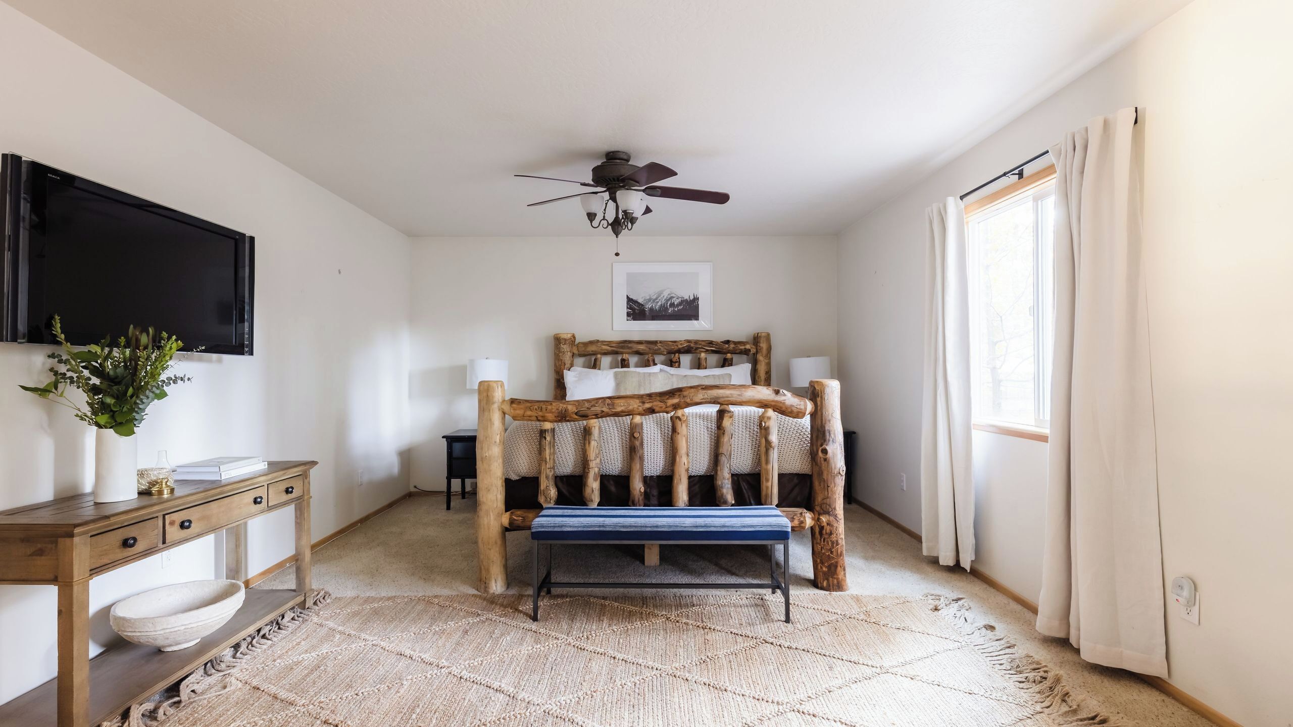 A bedroom with a wooden bed frame, a ceiling fan, a TV mounted on the wall, a desk with a plant, and a window with curtains.