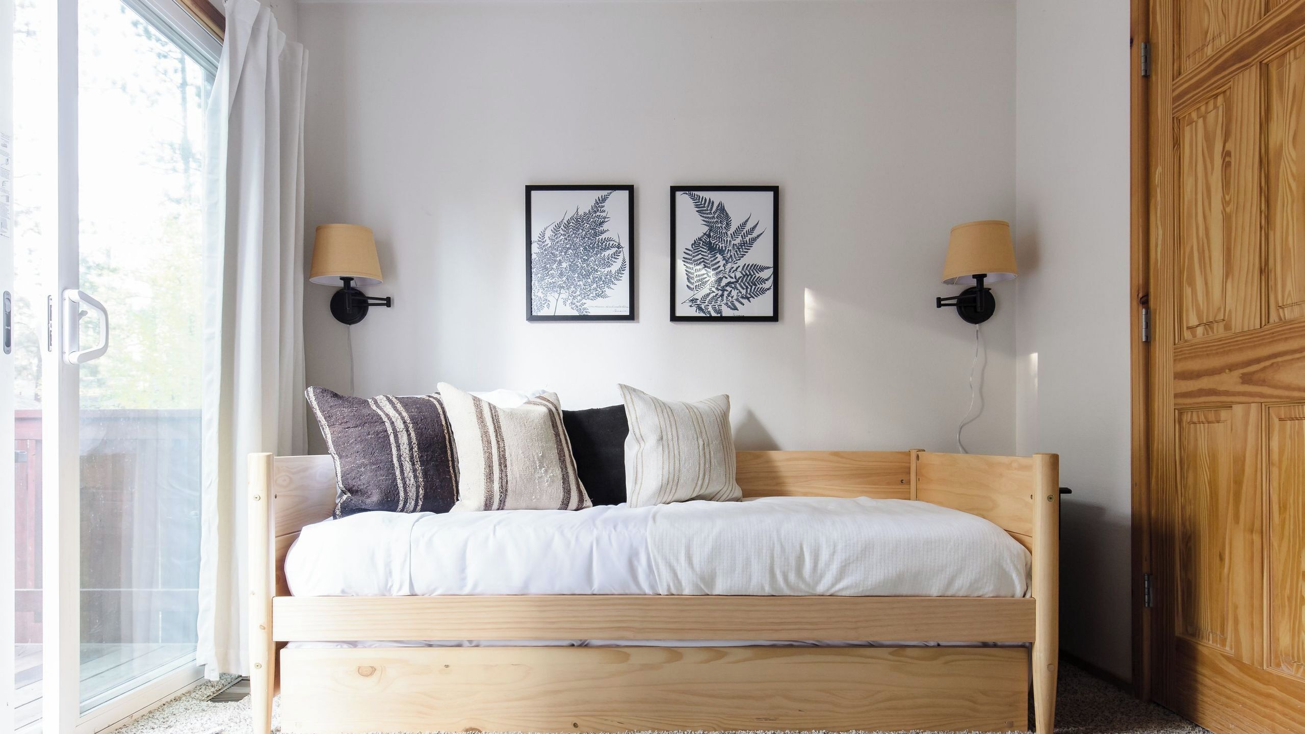Une chambre à coucher avec un lit en bois, du linge de lit blanc, deux lampes fixées au mur et deux reproductions de feuilles encadrées.