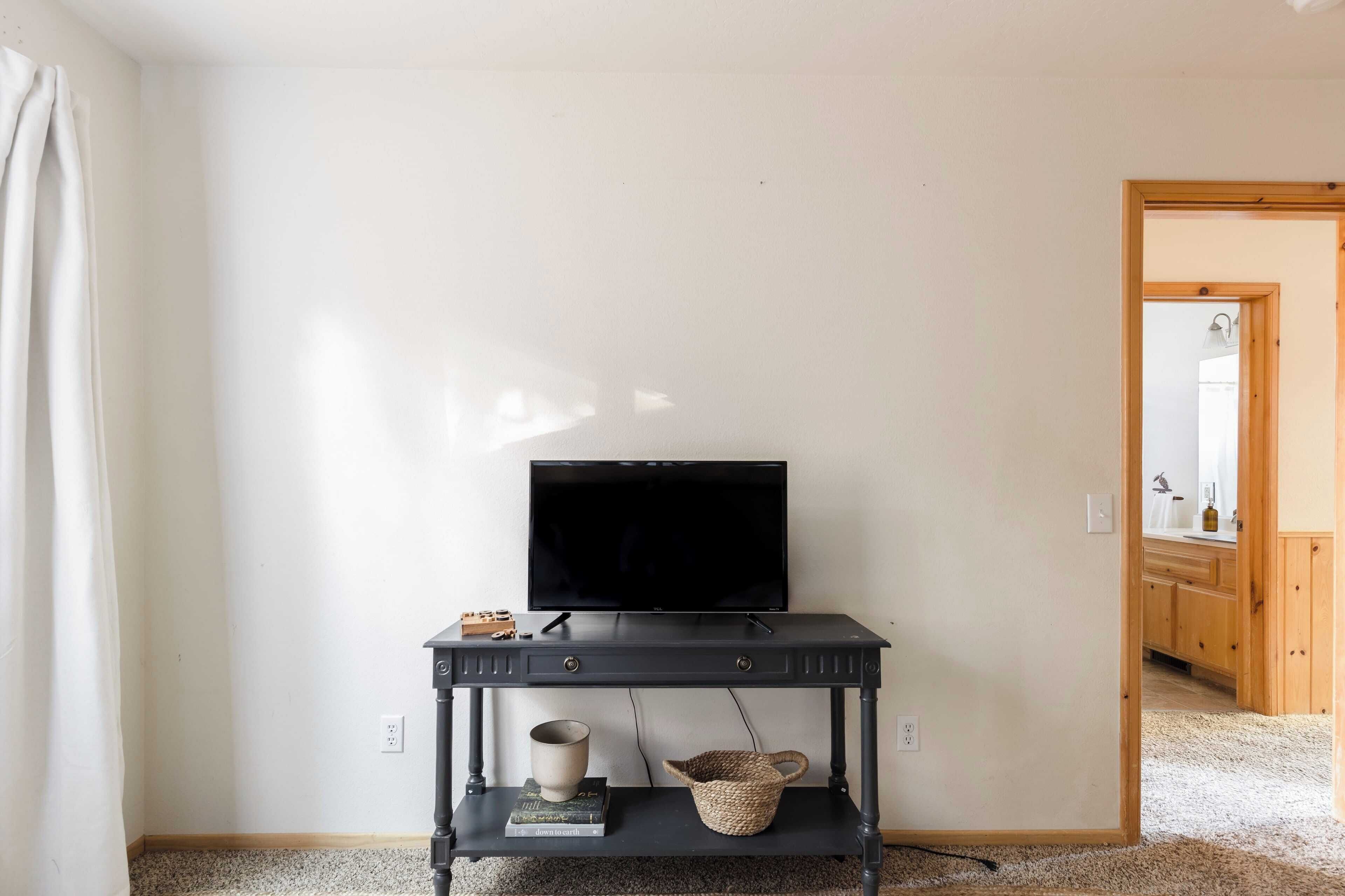 A room with a black TV on a stand, a small shelf with a cup and a basket, and a doorway leading to a bathroom.