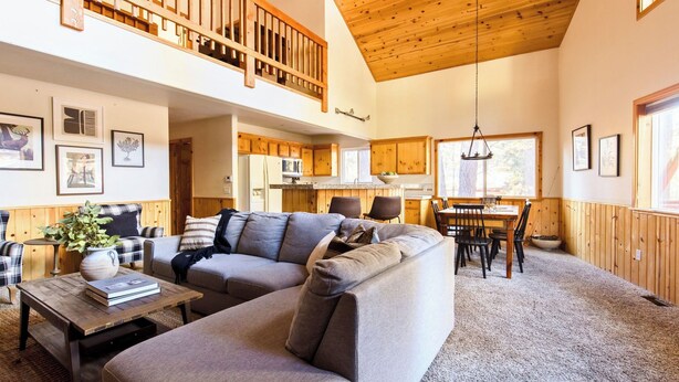 A spacious living area with a high ceiling, wooden beams, a dining table with chairs, and a kitchen in the background. - Walk to Ski Resort Lift Deck, Hot Tub, Game Loft Jumper by Avantstay (Big Bear Lake)