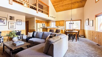 A spacious living area with a high ceiling, wooden beams, a dining table with chairs, and a kitchen in the background.