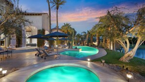 A pool area with lounge chairs and umbrellas, surrounded by palm trees and a building with arched windows.