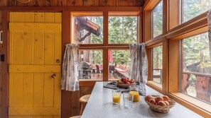 L’intérieur d’un chalet en bois, avec une table à manger, des aliments pour le petit-déjeuner et une vue sur une terrasse aménagée avec du mobilier d’extérieur.