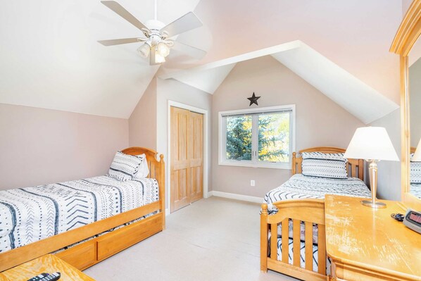 A bedroom with two single beds, a ceiling fan, a window with a view of trees, and a wooden dresser with a lamp.