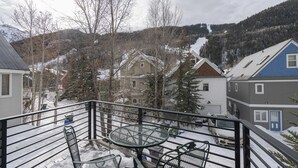 A balcony with a metal table and chairs, overlooking a snowy residential area with houses and trees.