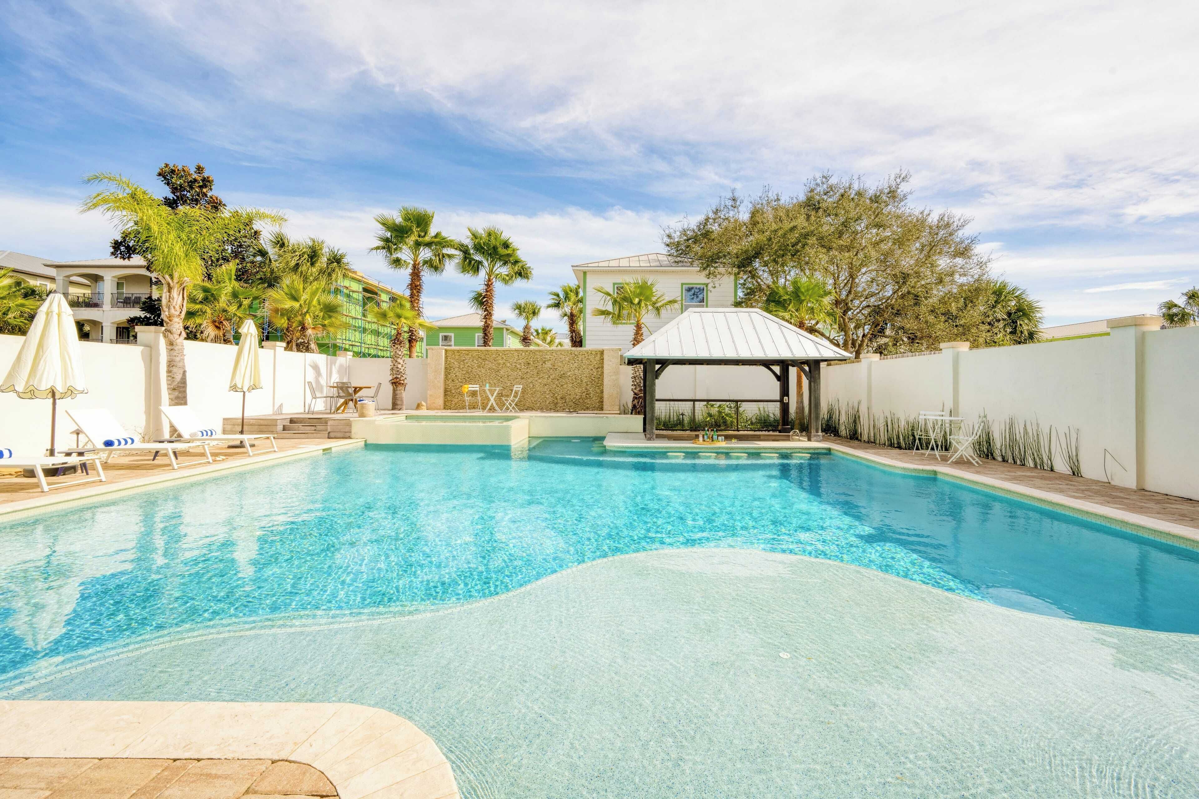A swimming pool with lounge chairs and umbrellas, surrounded by palm trees and a covered seating area.