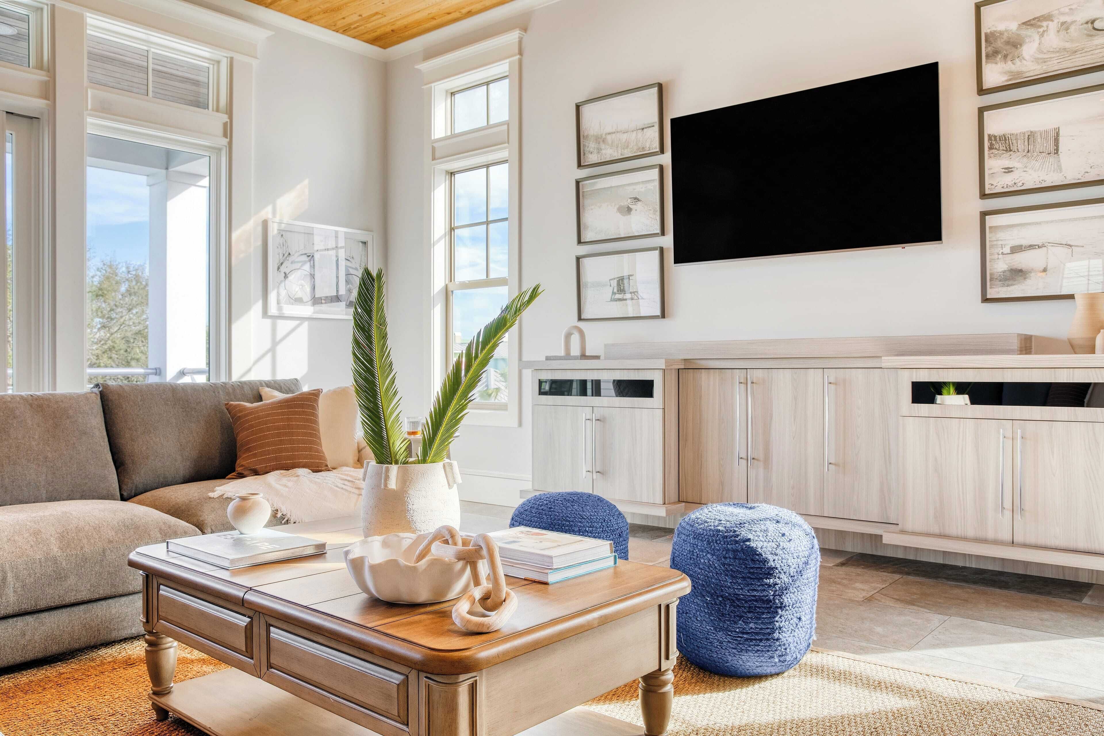 A living room with a sofa, coffee table, and blue pouf. There is a TV mounted on the wall and a window providing natural light.