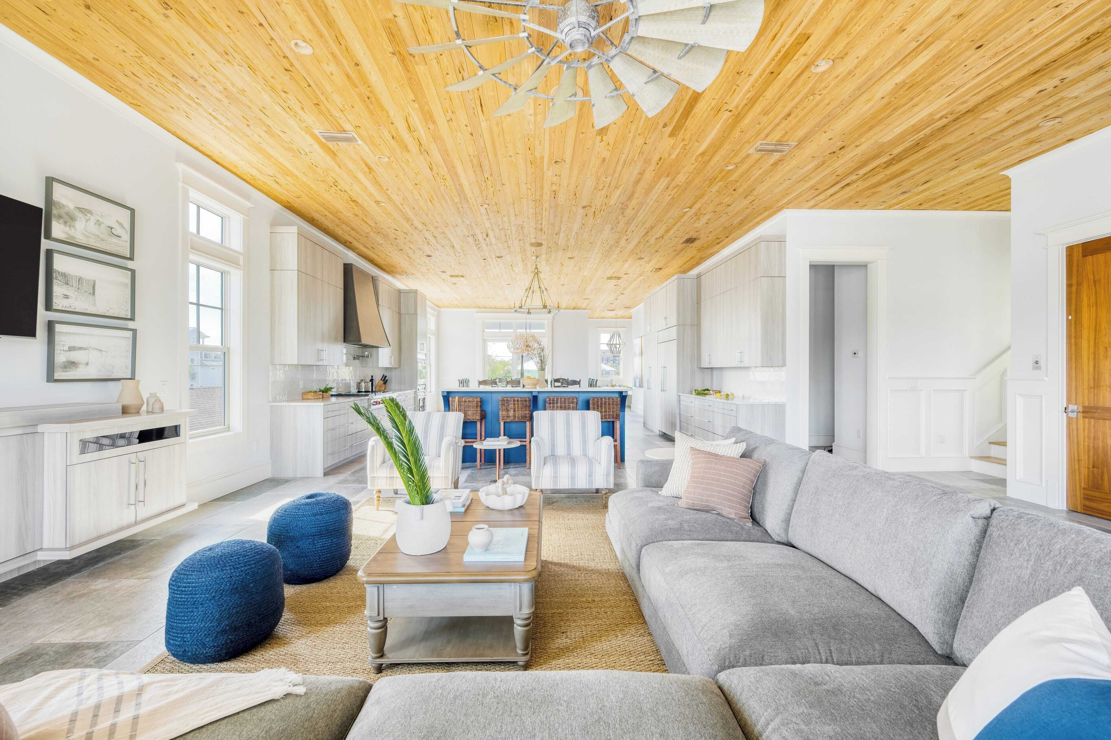 Un vaste espace de vie avec un plafond en bois, un canapé gris, des poufs bleus et une cuisine en arrière-plan.
