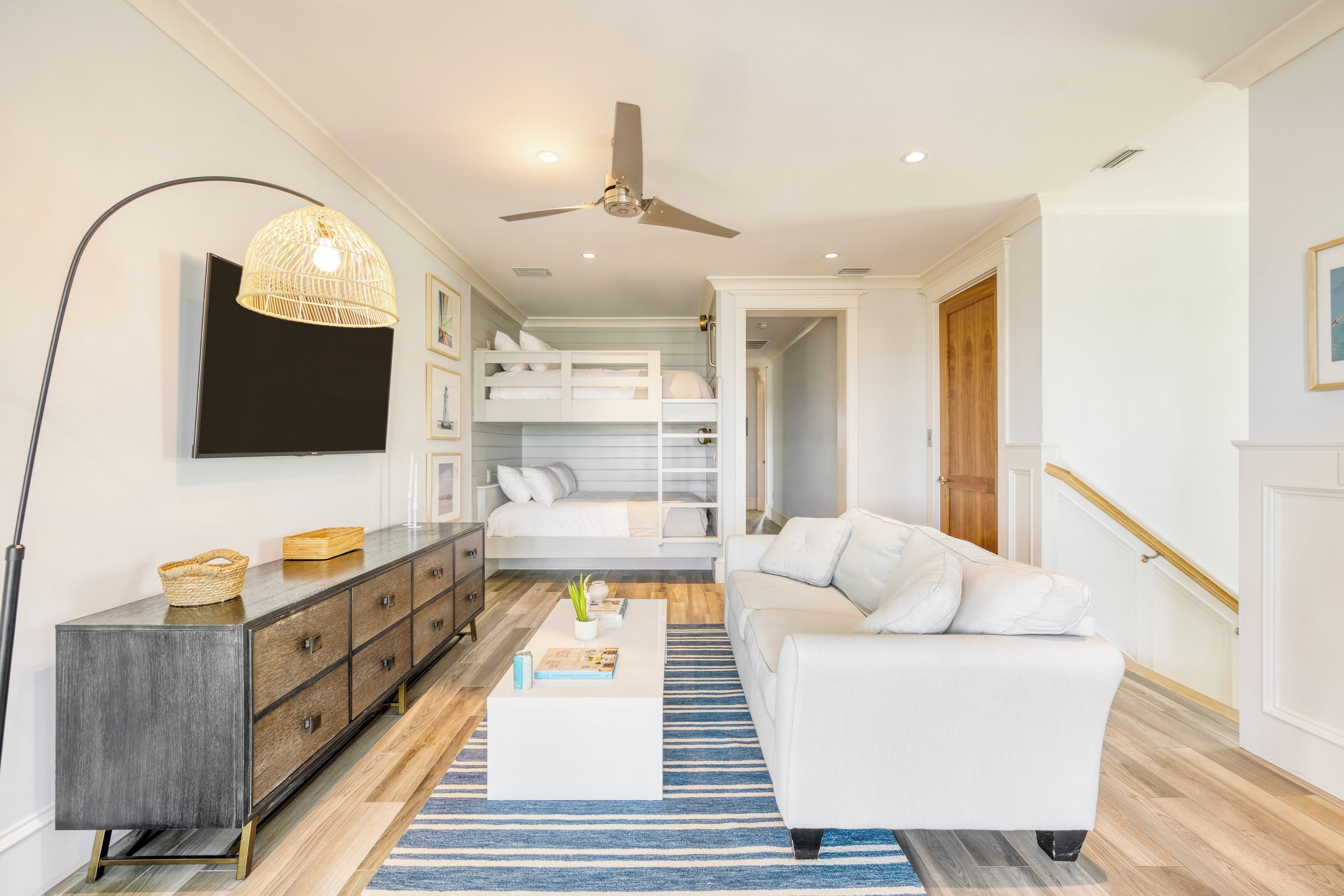 A modern living room with a white sofa, a coffee table, a dark wooden cabinet, a TV mounted on the wall, and a ceiling fan.