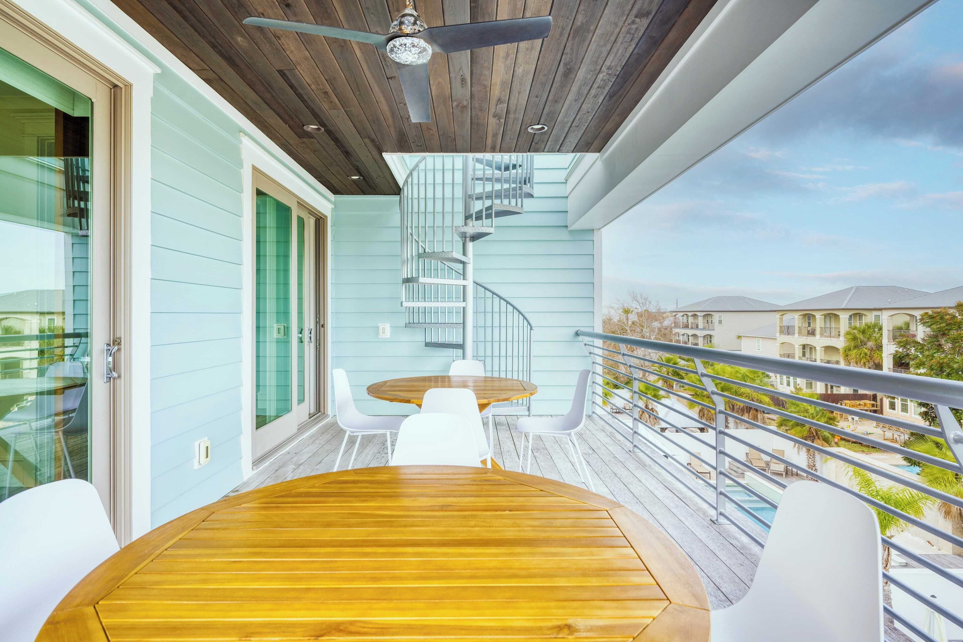 Une terrasse en bois avec une table ronde et des chaises blanches, un escalier en colimaçon et une vue sur une piscine et des bâtiments en arrière-plan.