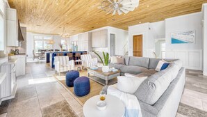 A spacious living area with a wooden ceiling, a gray sofa, and a dining area in the background. - Monarch by Avantstay Stunning Estate 1 Block to Beach, Swim Up Bar, Hot Tub, & Rooftop Views (Destin)