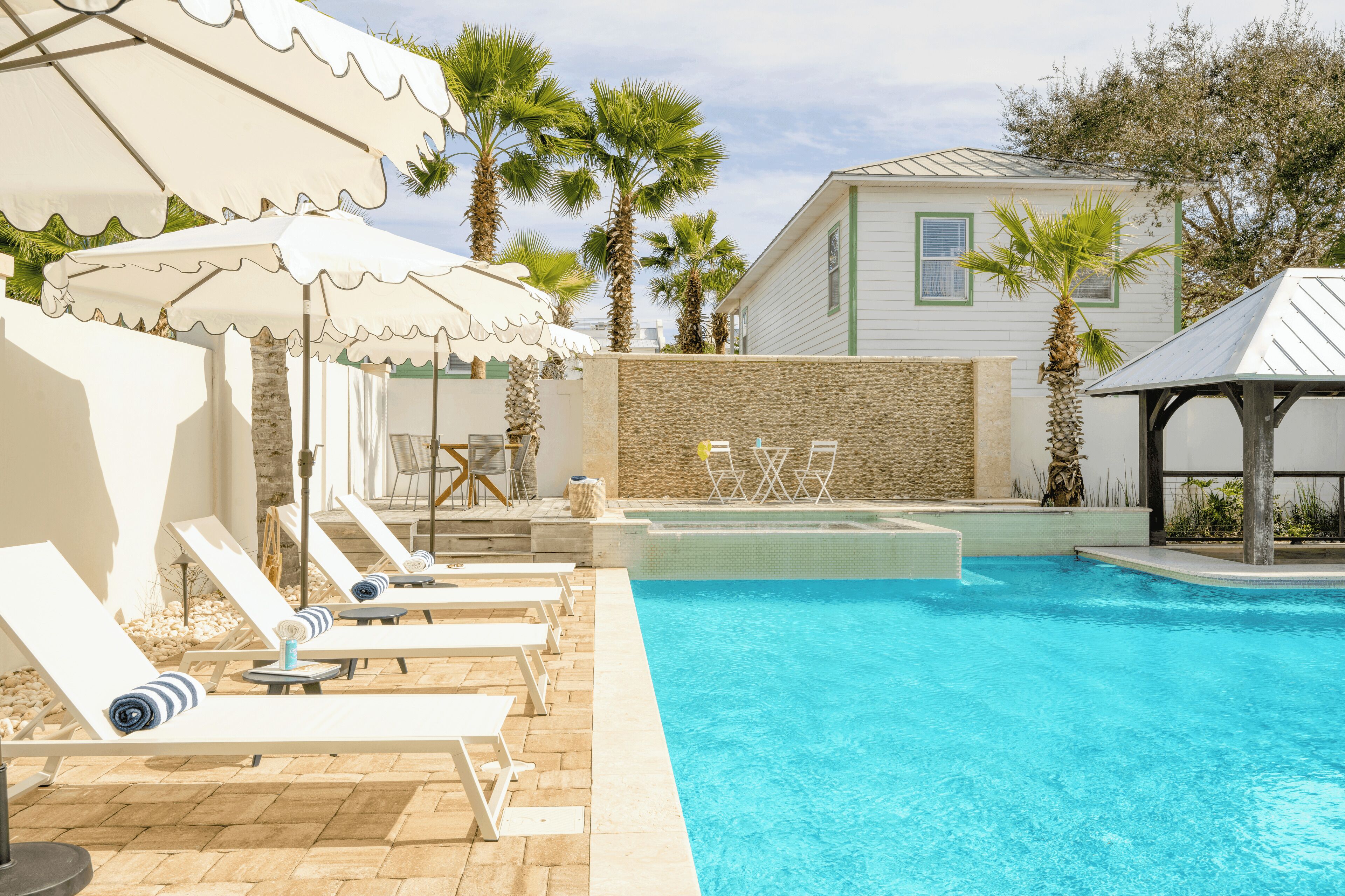 Un espace piscine aménagé avec des chaises longues et des parasols, un mur en pierre et des palmiers.