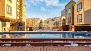 A swimming pool with wooden steps leading up to it, surrounded by buildings with stone and wood exteriors, and a view of mountains in the background. - Cloud 9 by Avantstay Located at the Base of Vail Ski Resort w/ Community Pool & Hot Tub (Vail)