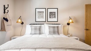 A neatly made bed with a white duvet and two pillows, a bedside lamp, a framed picture on the wall, and a wooden door.