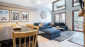 A modern living room with a dining table, blue sofa, and large windows. - Cloud 9 by Avantstay Located at the Base of Vail Ski Resort w/ Community Pool & Hot Tub (Vail)