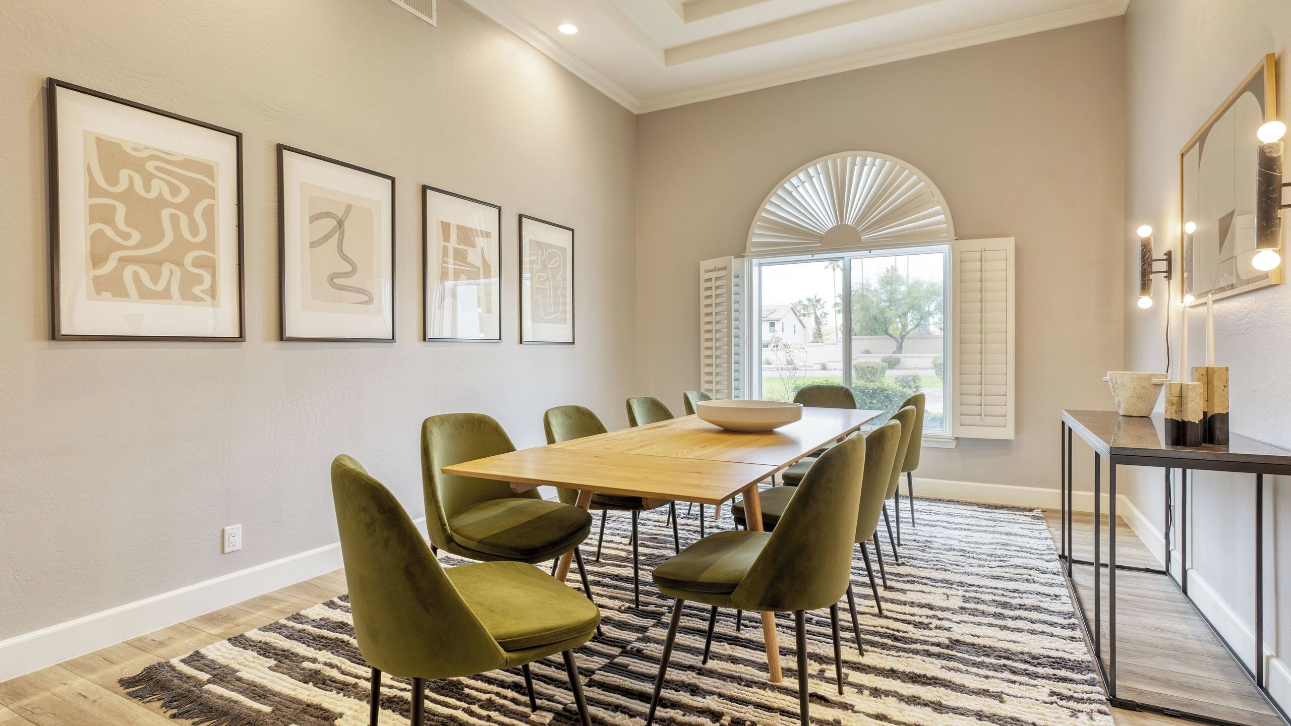 A dining area with a wooden table and green chairs, framed artwork on the wall, and a large window with shutters.