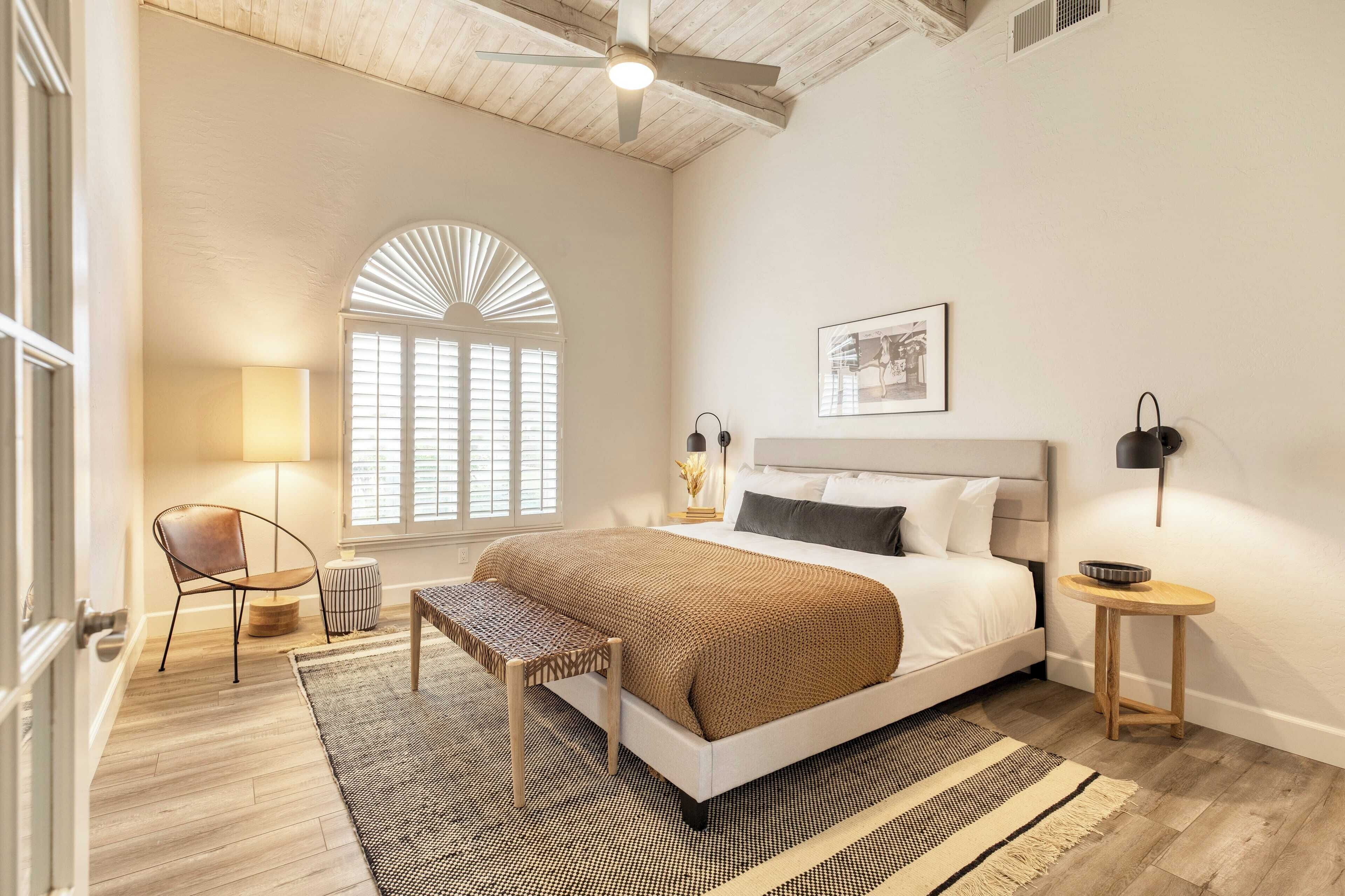 Une chambre à coucher comprenant un lit, une chaise, une lampe, une table de chevet et une fenêtre avec des stores.
