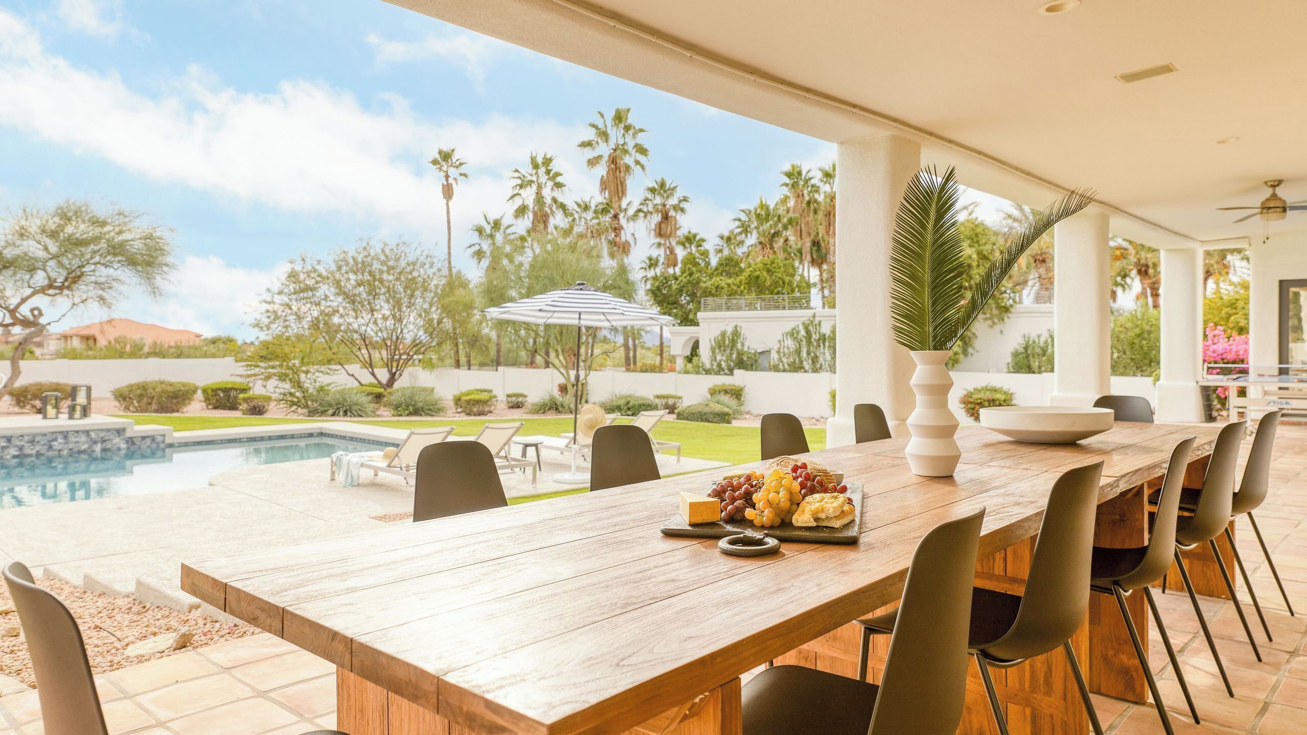 An outdoor dining area with a wooden table, chairs, a bowl of fruit, and a view of a swimming pool and palm trees.