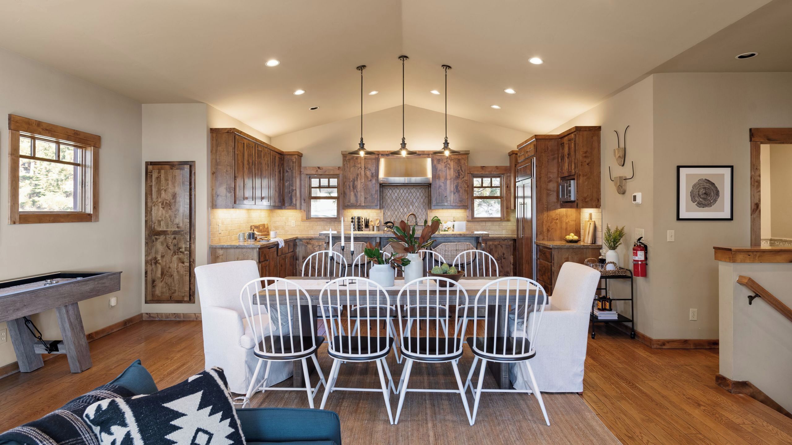 A modern kitchen with wooden cabinets, a dining area with white chairs, and a pool table.