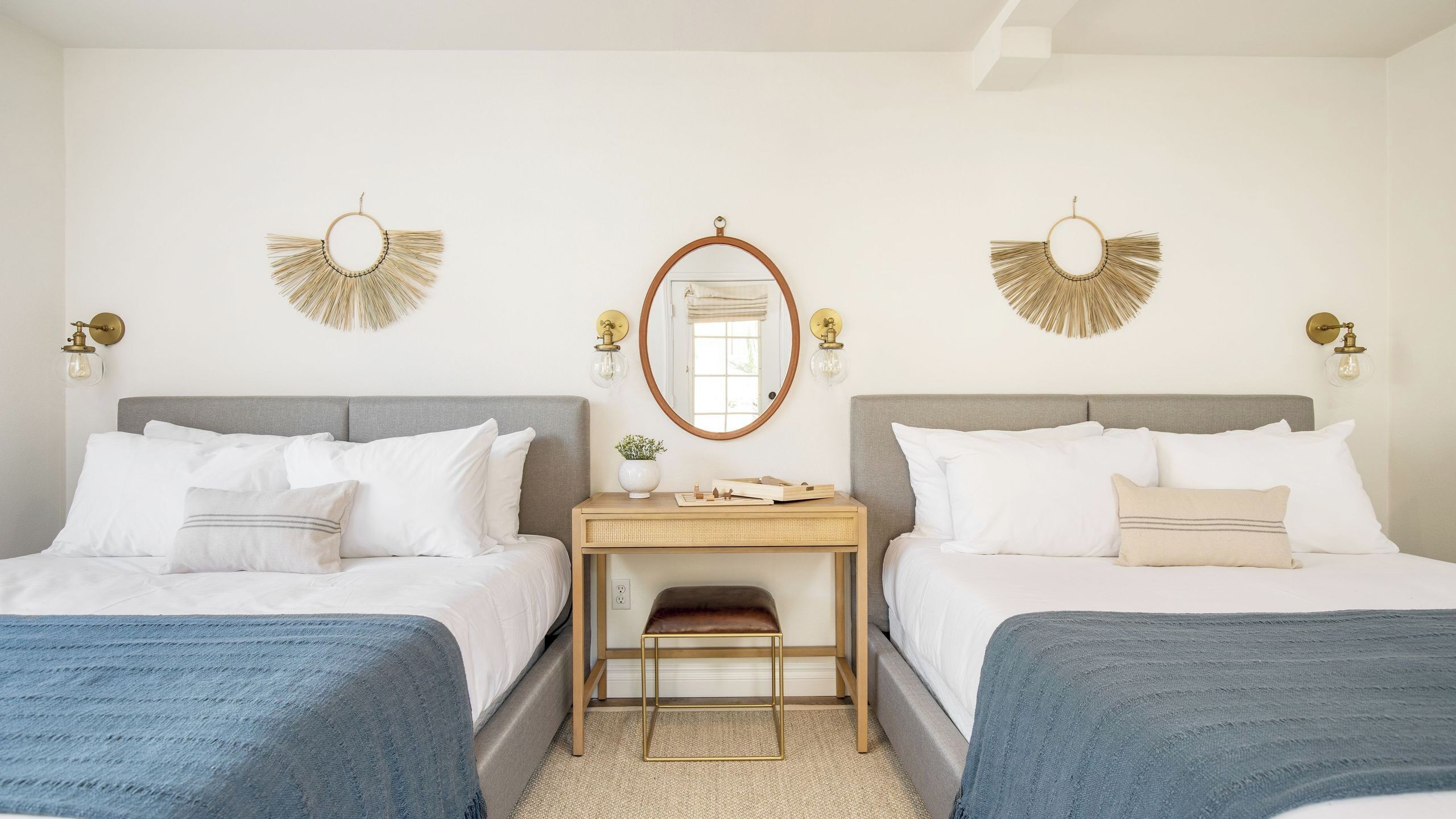 A hotel room with two beds, a small desk, a mirror, and wall sconces.