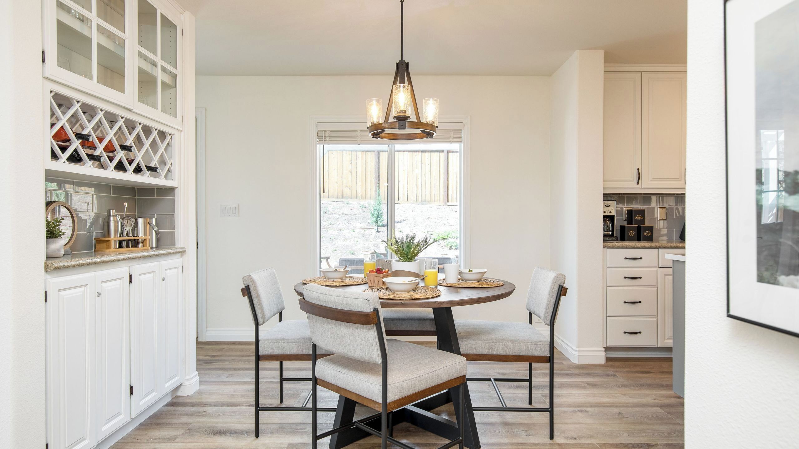 A dining area with a round table, four chairs, a chandelier, and a view through a window.