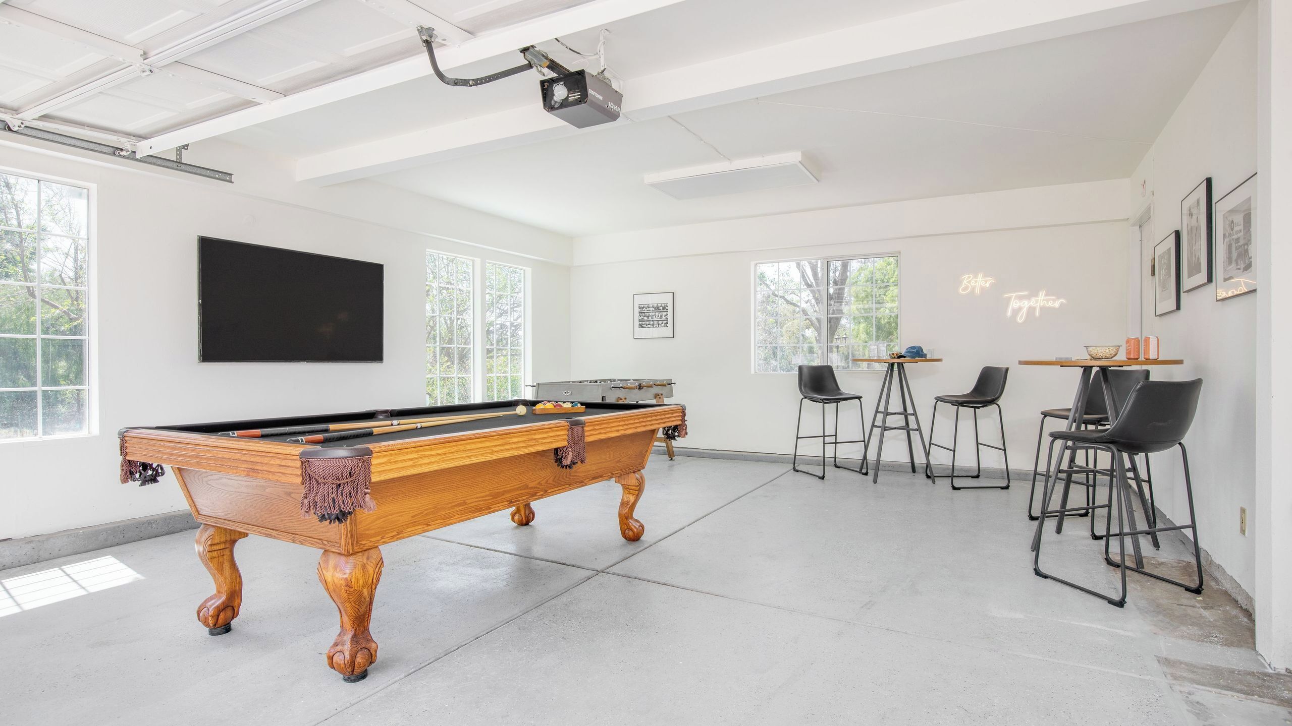 A pool table in a bright room with large windows, a projector, and bar stools.