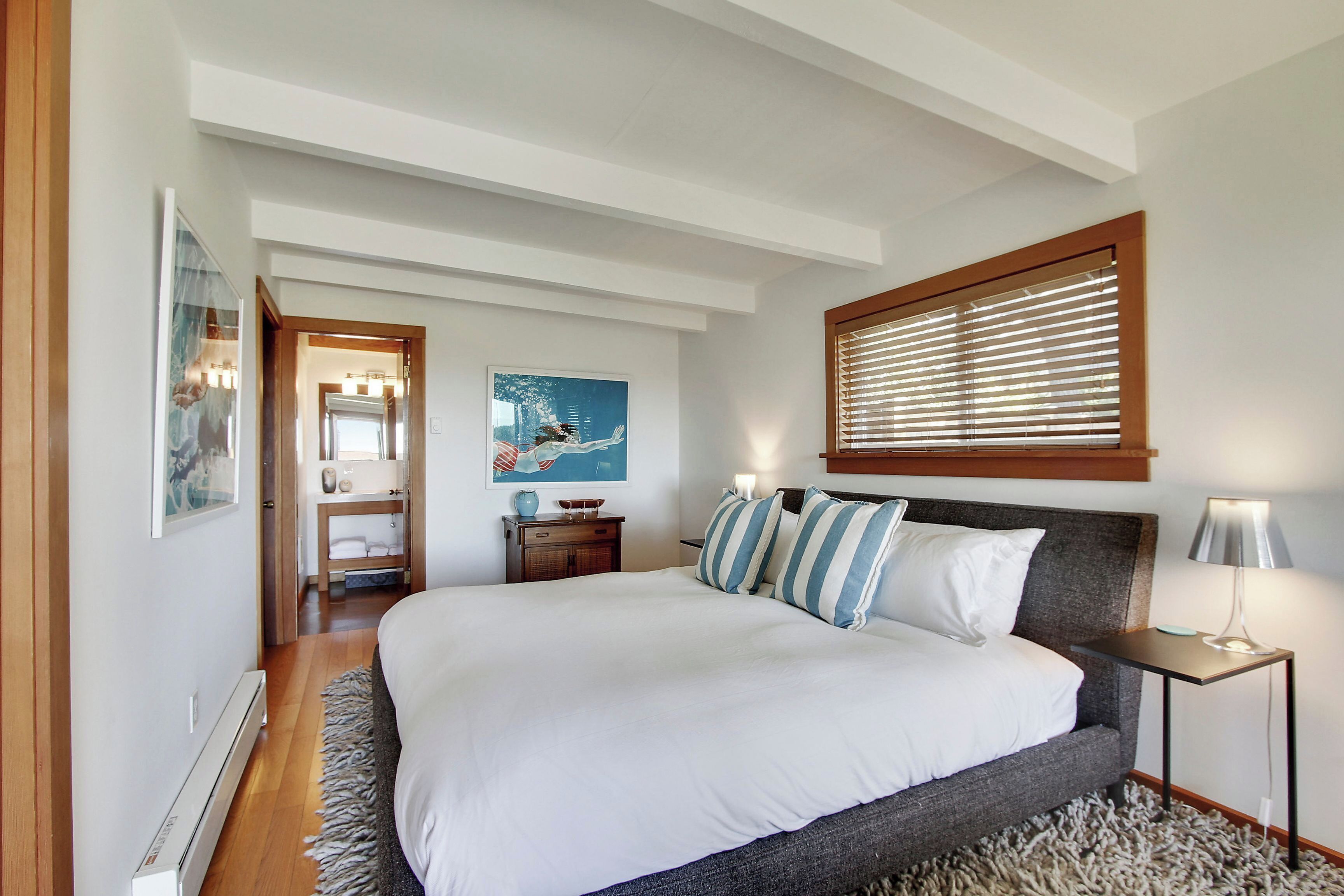 A bedroom with a large bed, a bedside table with a lamp, a window with blinds, a door leading to a bathroom, and a painting on the wall.