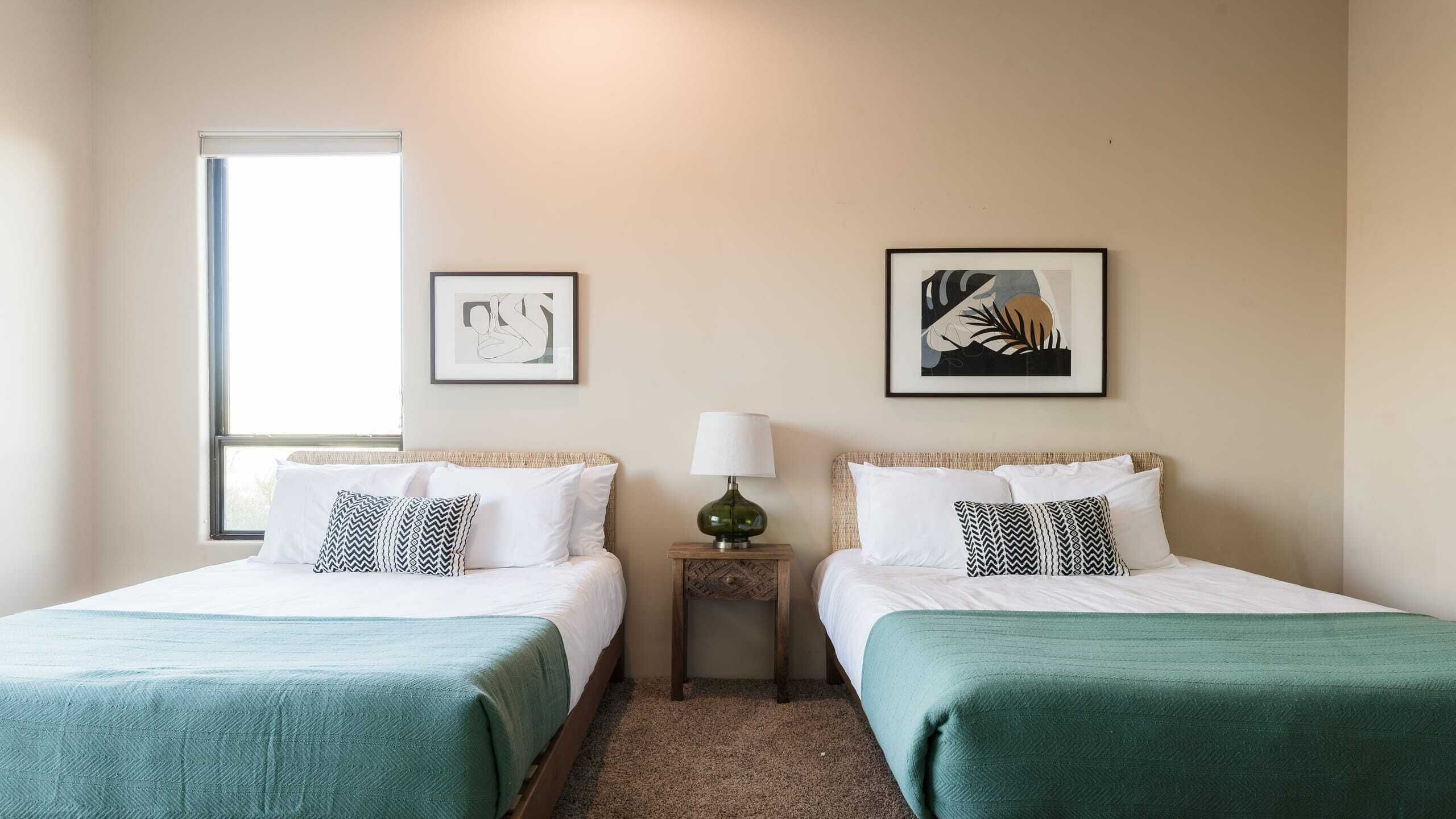 A hotel room with two single beds, a small table with a lamp, and two framed artworks on the wall.